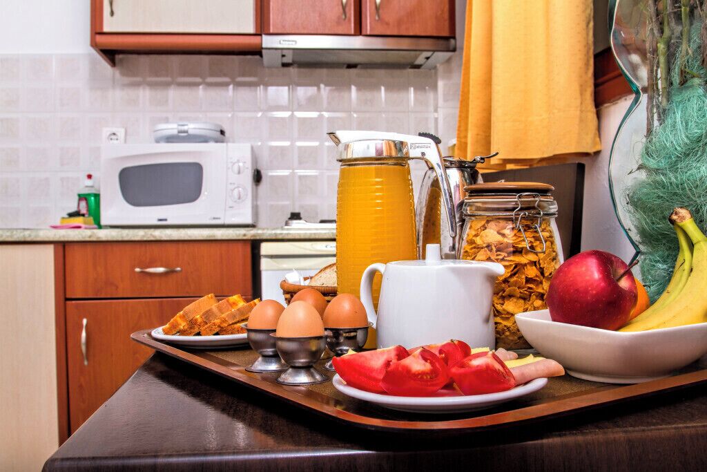 A kitchen counter with a microwave, a coffee maker, a jar of cereal, eggs, a plate of toast, a bowl of fruit, and a cutting board with tomatoes.