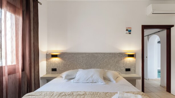 A hotel room with a bed, two bedside tables, a headboard, and a view of the outside. - Birds Villa Apartments with Heated Pool - Including Car and Breakfast (Santorini)