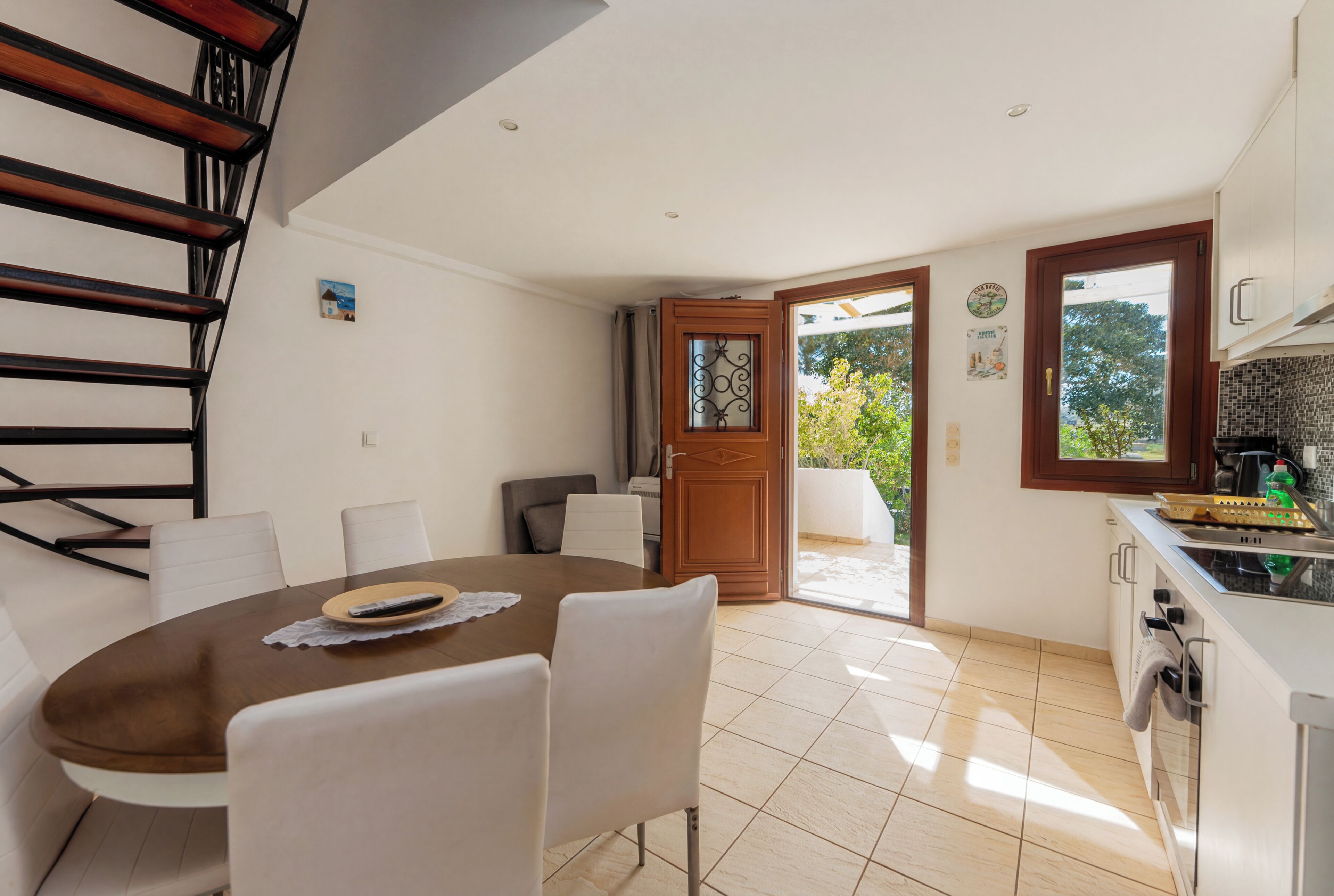 A dining area with a round table, chairs, a staircase, and a kitchen with a window.