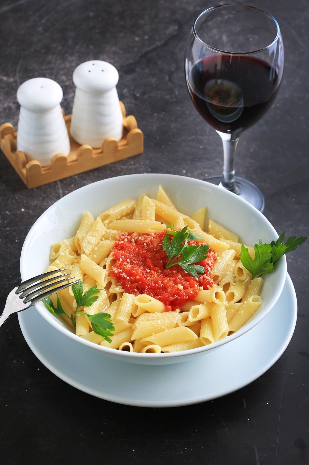 A bowl of pasta with a red sauce garnish, a fork, and a glass of red wine on a dark surface.