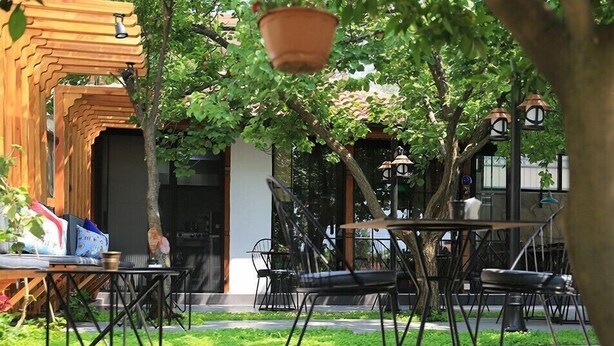 Un área de estar al aire libre con mesas y sillas, rodeada de vegetación y un edificio con fachada de madera.