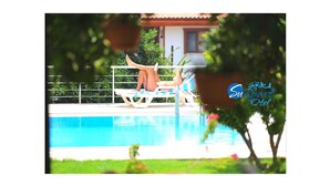 Zona de piscina con una persona descansando en una tumbona, una valla y un edificio al fondo.