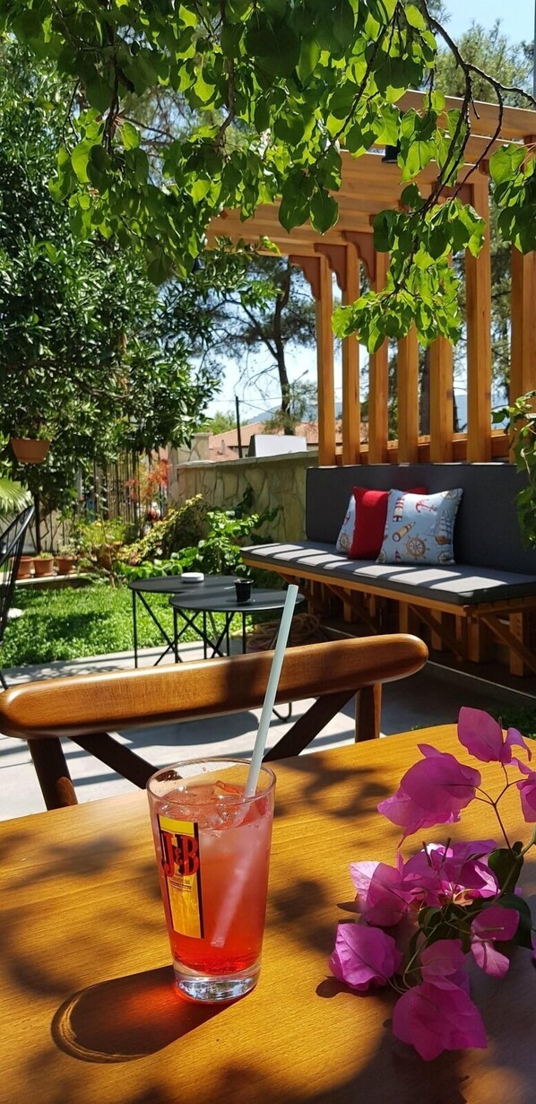 Una mesa de madera con una bebida y una pajita, rodeada de follaje y una pérgola de madera.