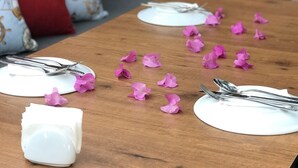 Una mesa de comedor de madera dispuesta con platos, cubiertos y pétalos de flores rosados.