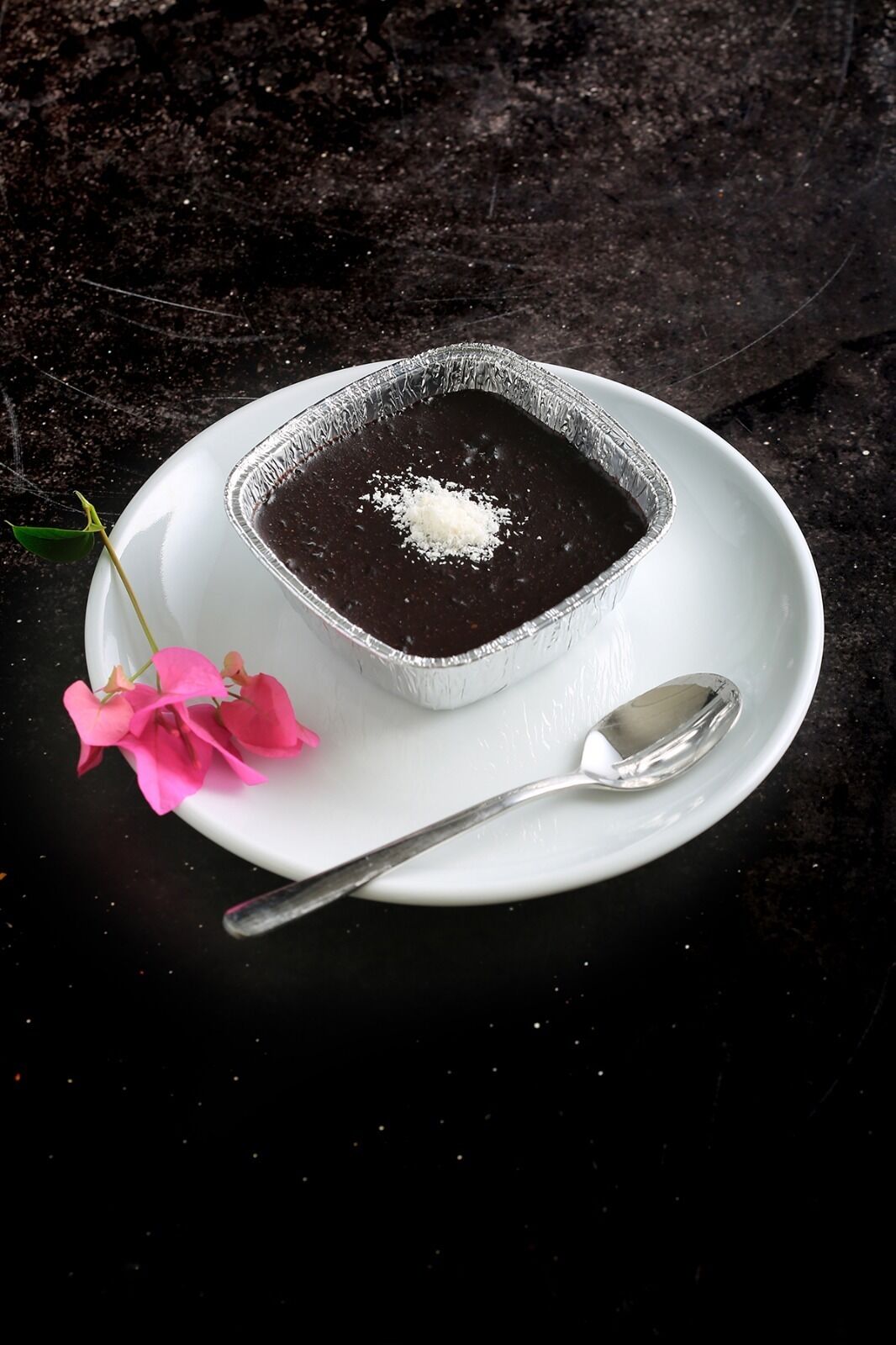 A dessert in a foil dish on a white plate with a spoon, accompanied by pink flowers.