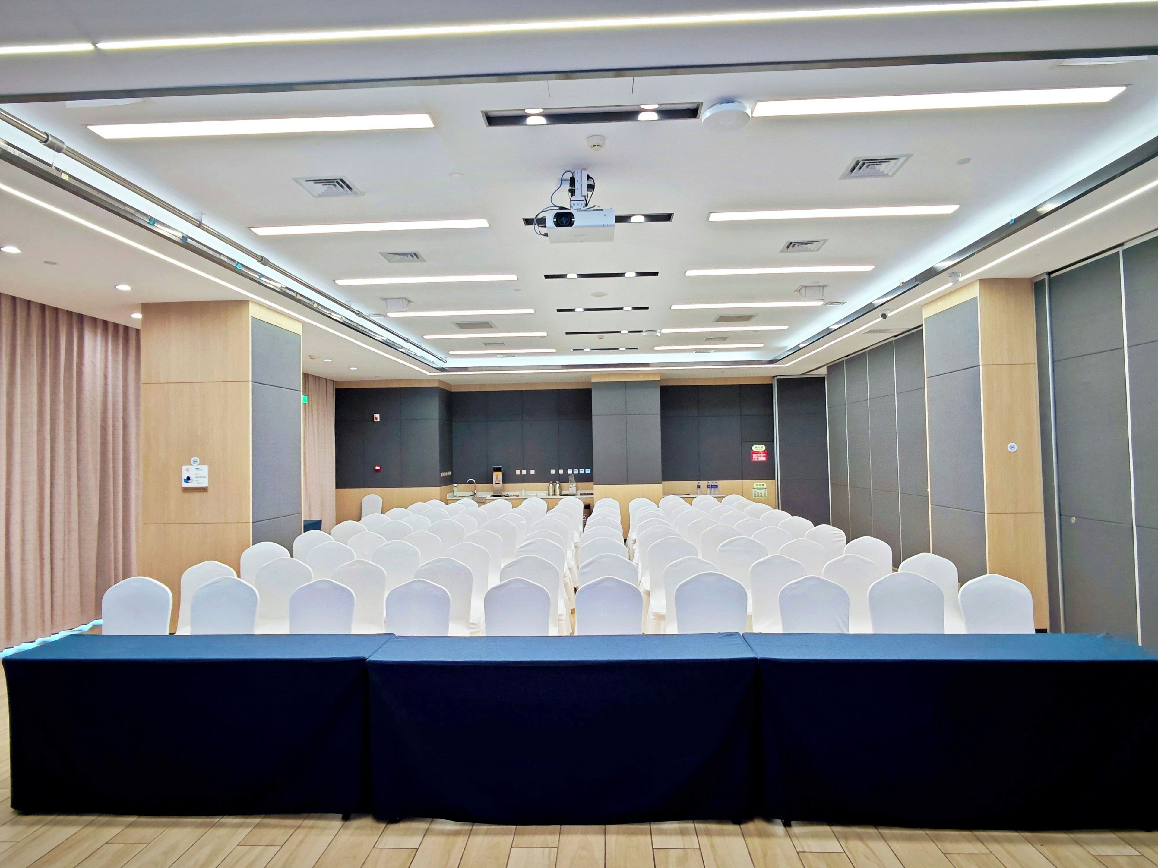 Un salón de conferencias con filas de sillas blancas, una mesa larga y un proyector en el techo.