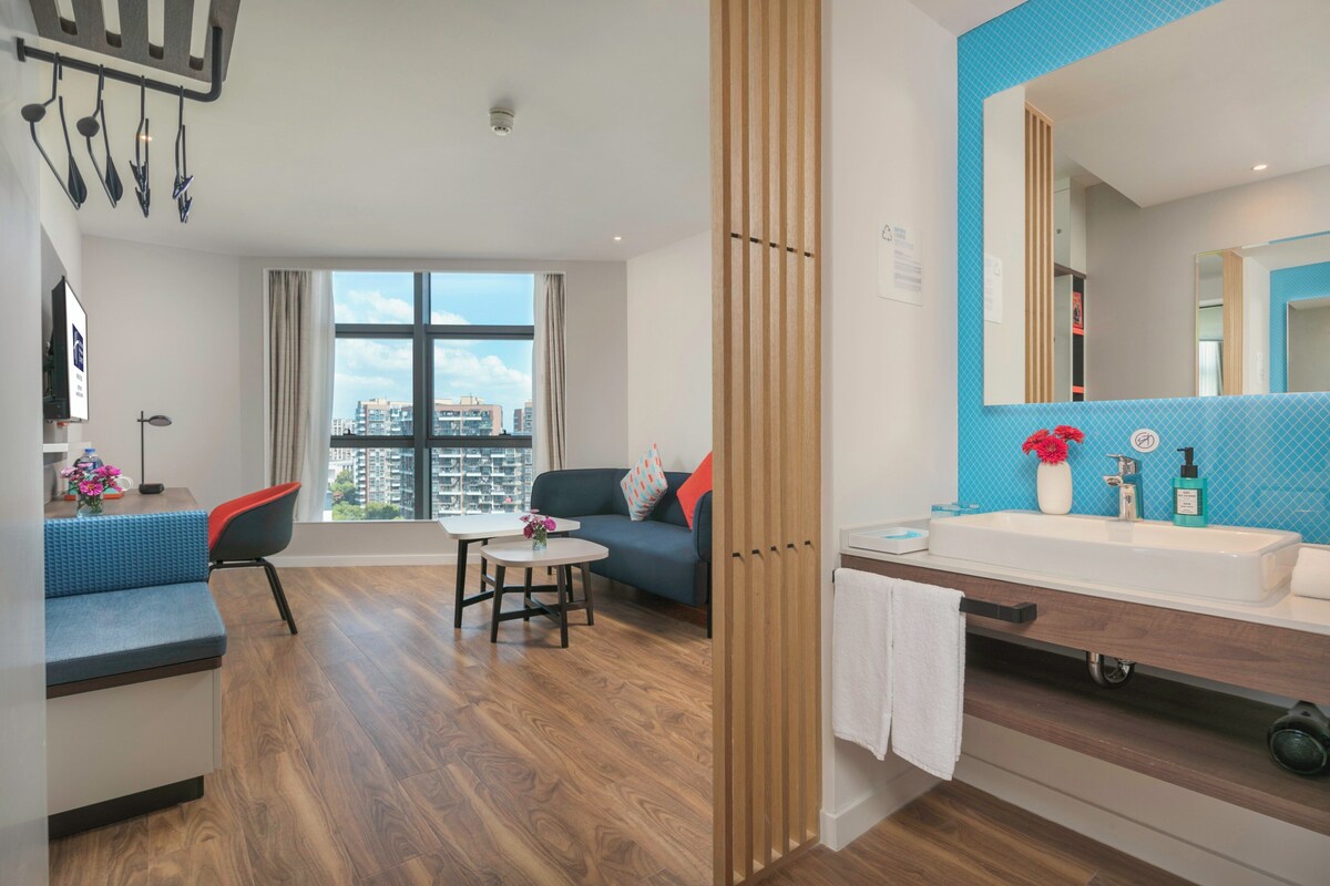 A modern hotel room with a large window, a sofa, a small table, a bathroom with a sink and mirror, and a city view.