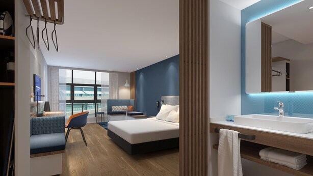 A modern hotel room with a large bed, a seating area with a chair and table, a large window, and a bathroom with a sink and mirror. - Holiday Inn Express Hangzhou Jiuzhou by IHG (Hangzhou)