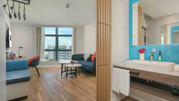 Una habitación de hotel moderna con un ventanal grande, un sofá, una mesita pequeña, un armario de madera y un baño con lavamanos y espejo.
