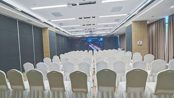 Een congreszaal met rijen witte stoelen, een podium met een groot scherm en een houten vloer.