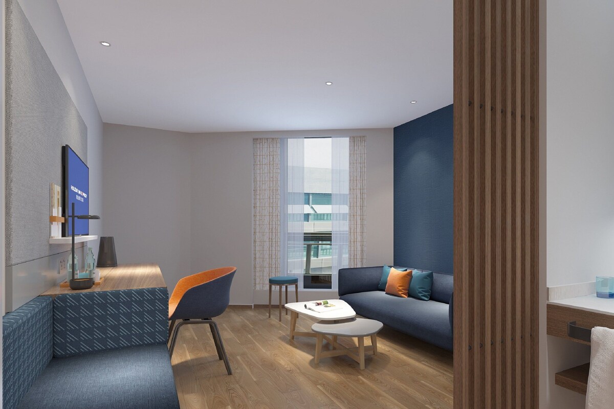 Una habitación de hotel moderna con sofá, una mesa de centro, televisor y baño con espejo y lavamanos.