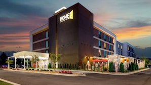 Un moderno edificio de hotel con un llamativo letrero que dice "HOME 2", un espacio exterior cubierto con asientos y una entrada ajardinada.
