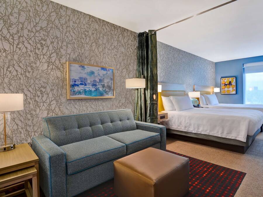 A hotel room with a grey sofa, a bed with white bedding, a small table, a lamp, and a cityscape painting on the wall.