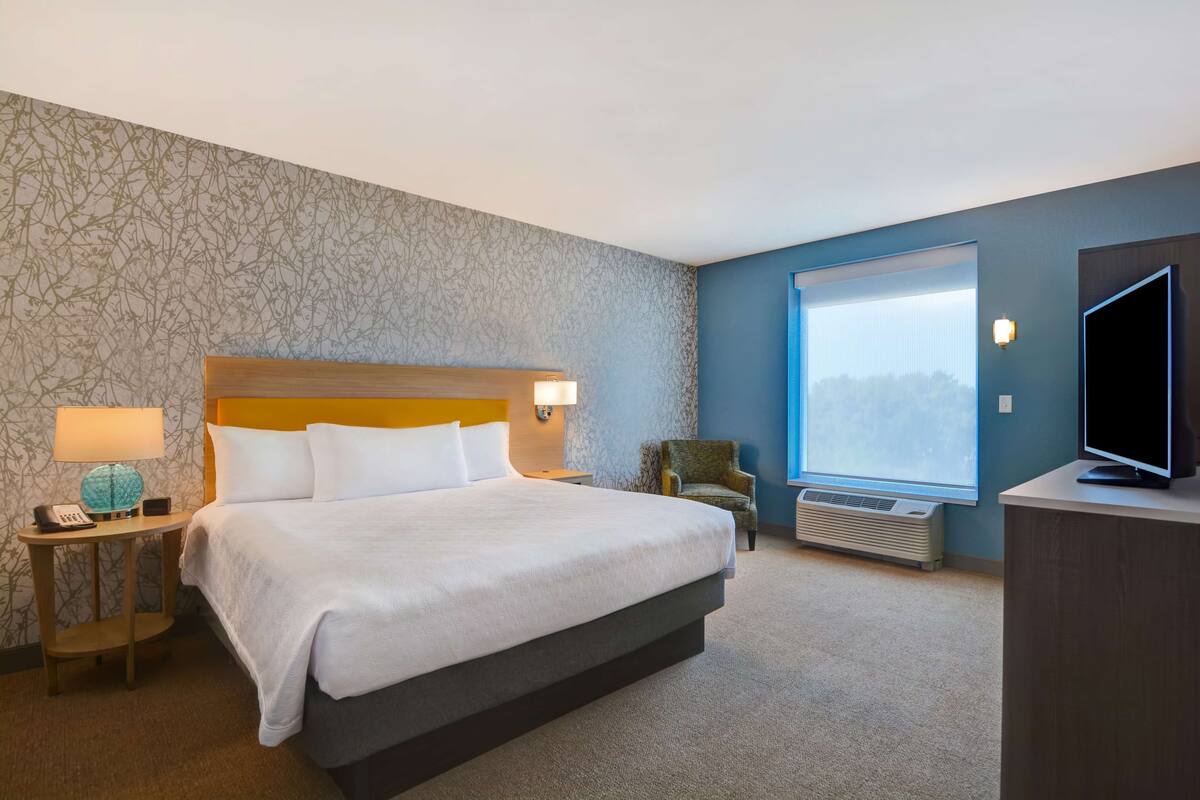 A hotel room with a large bed, a nightstand with a lamp, a chair, a TV on a stand, and a window with a view.