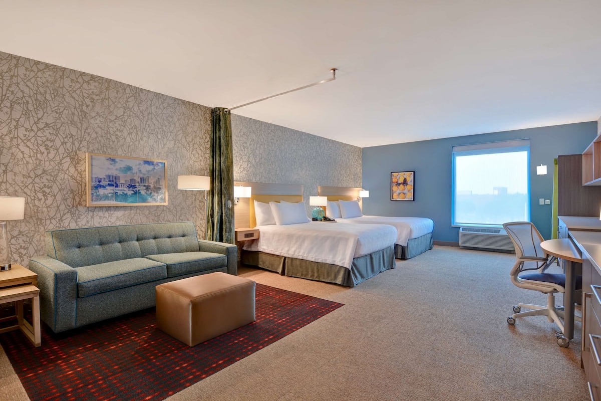 A hotel room with a sofa, two beds, a desk, and a view of the city.