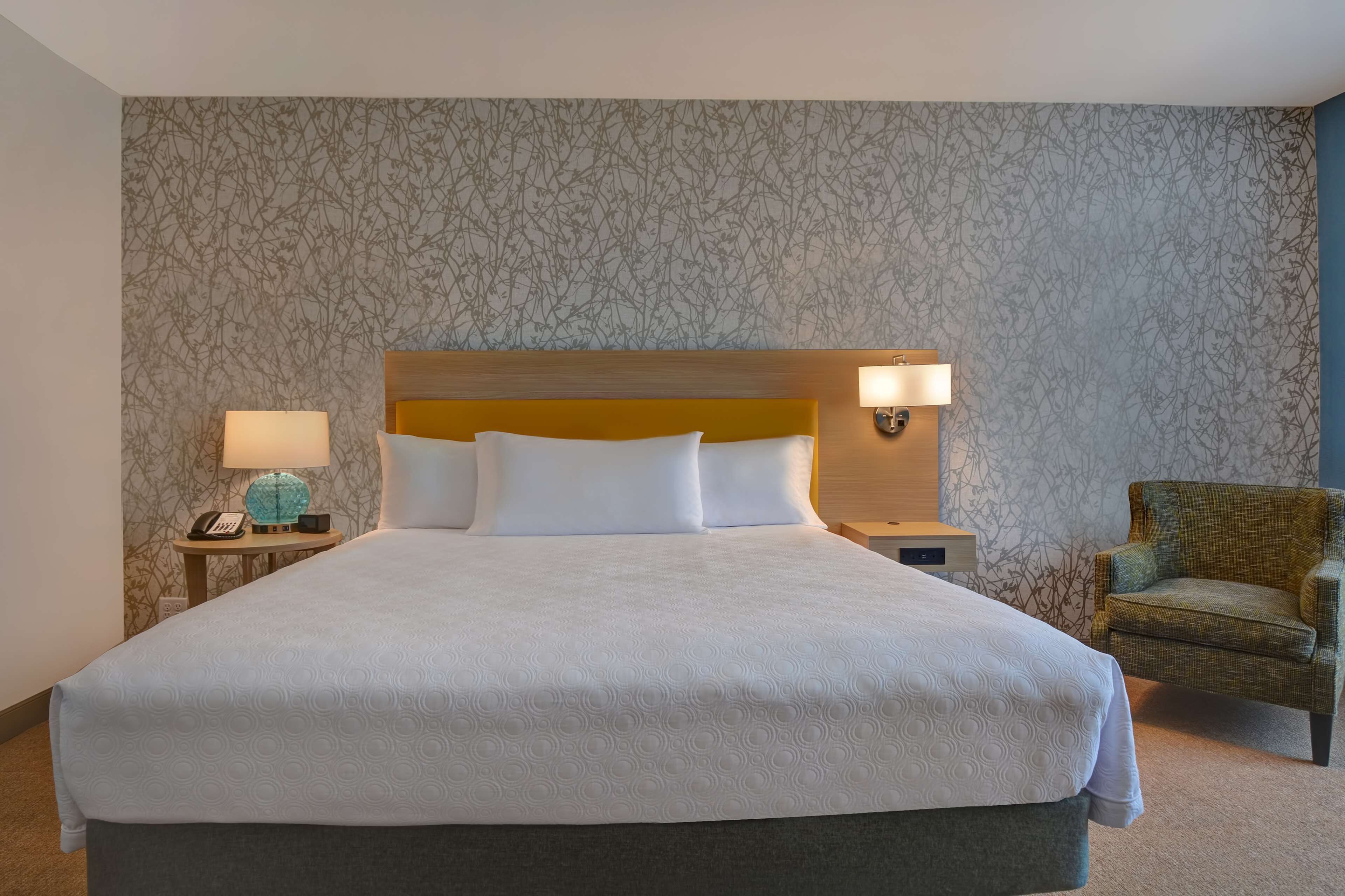 Een hotelkamer met een groot bed, twee bedlampen, een stoel en een behang met een patroon.