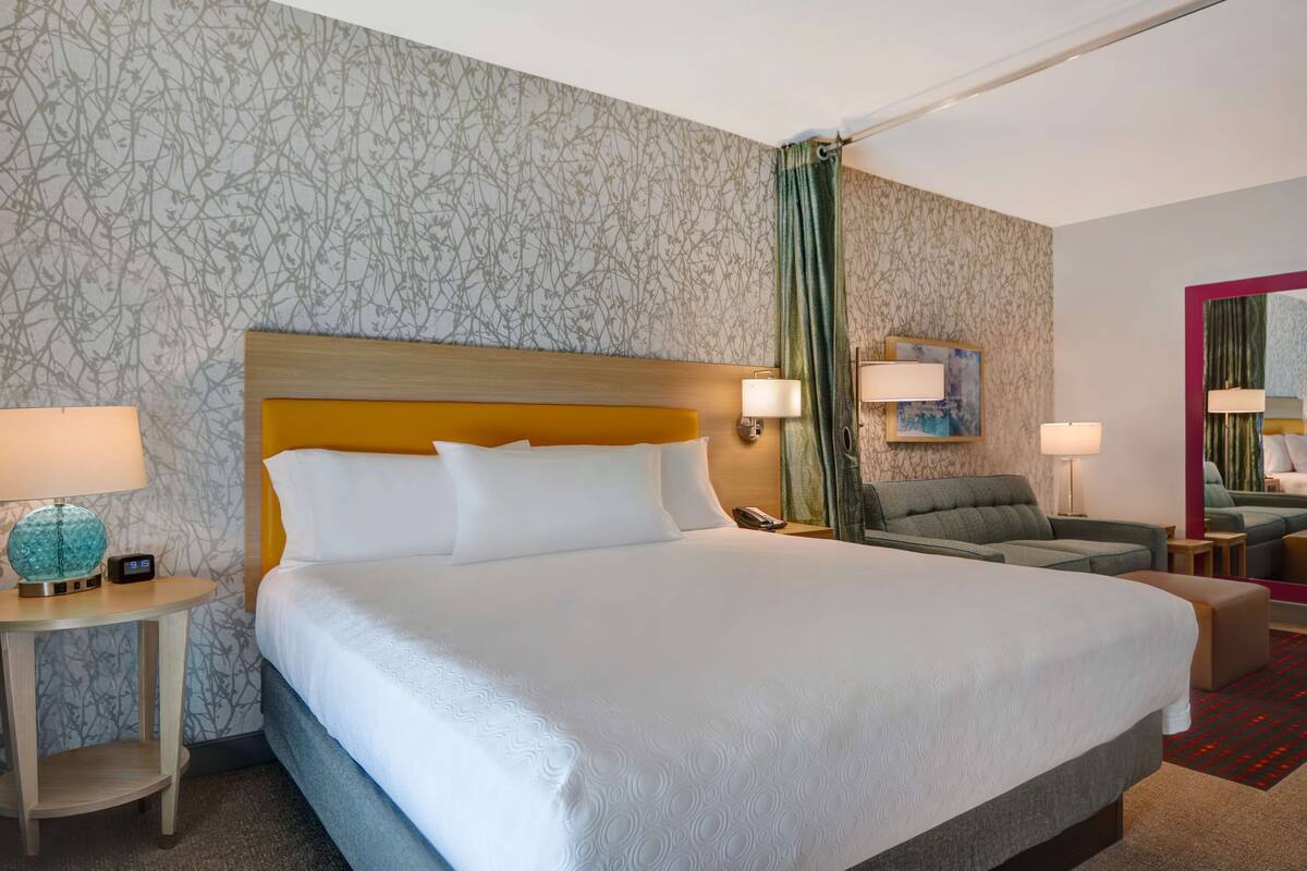 A hotel room with a large bed, a wooden headboard, a side table with a lamp, a green chair, and a small ottoman.