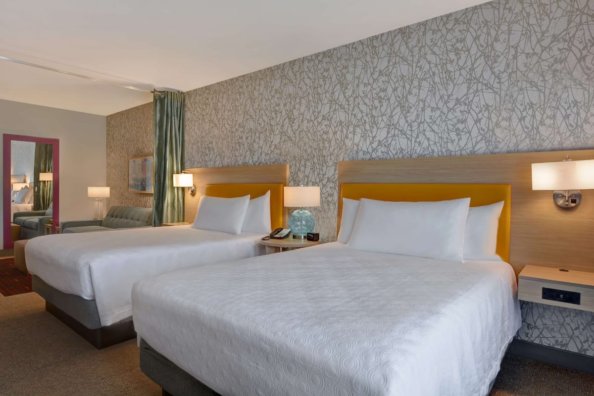 A hotel room with two beds, a small table with a lamp, and a chair.