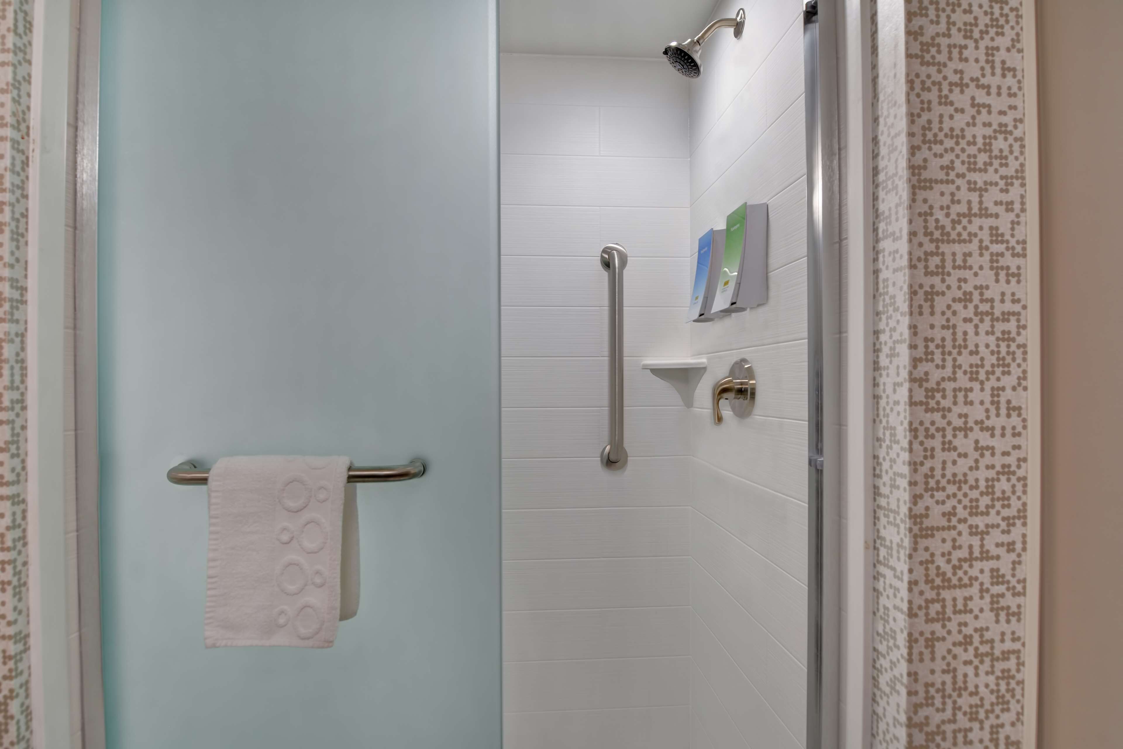 Un baño con paredes de azulejos blancos, cabina de ducha de vidrio, un perchero con una toalla blanca y un estante pequeño con artículos de aseo.