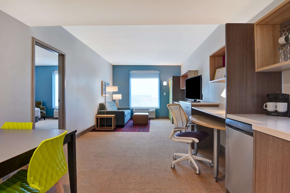 A hotel room with a dining area, a sofa, a desk with a computer, and a kitchen area with a coffee maker.