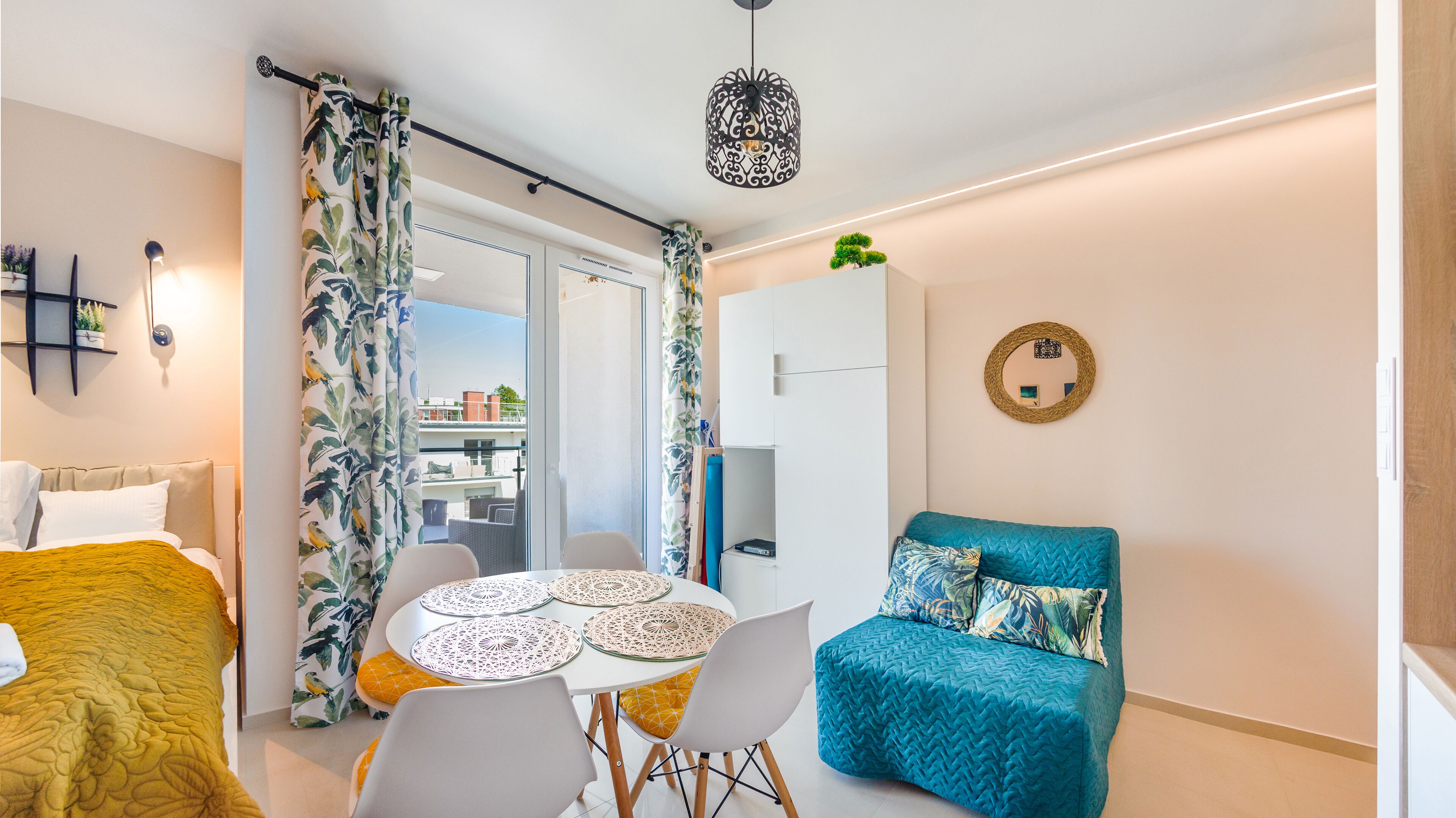 A compact living space with a bed, a sofa, a dining table with chairs, and a balcony with a view.
