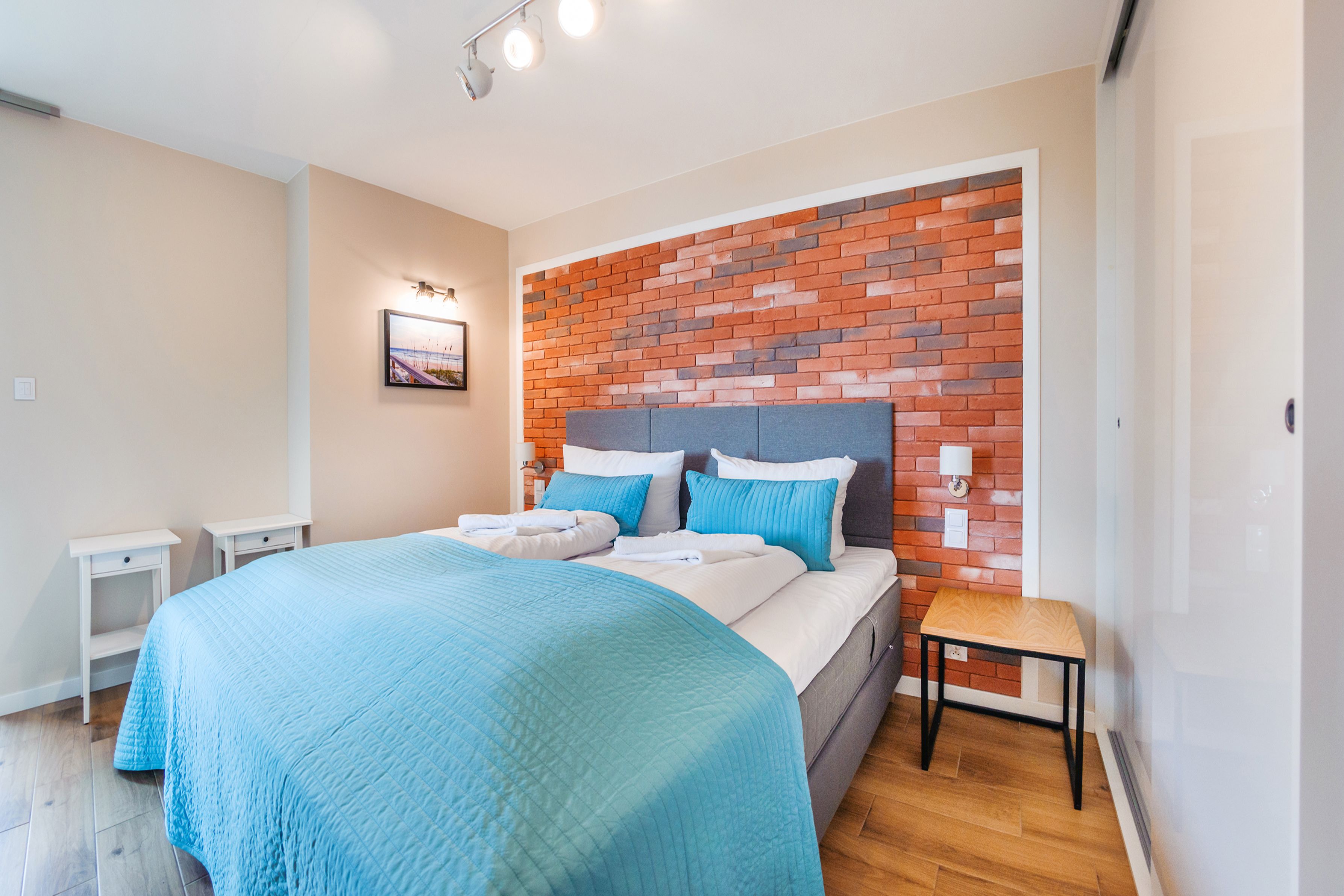 Une chambre à coucher comprenant un lit, une table de chevet, une petite table, une tête de lit ornée d’un motif en briques et un tableau accroché au mur.
