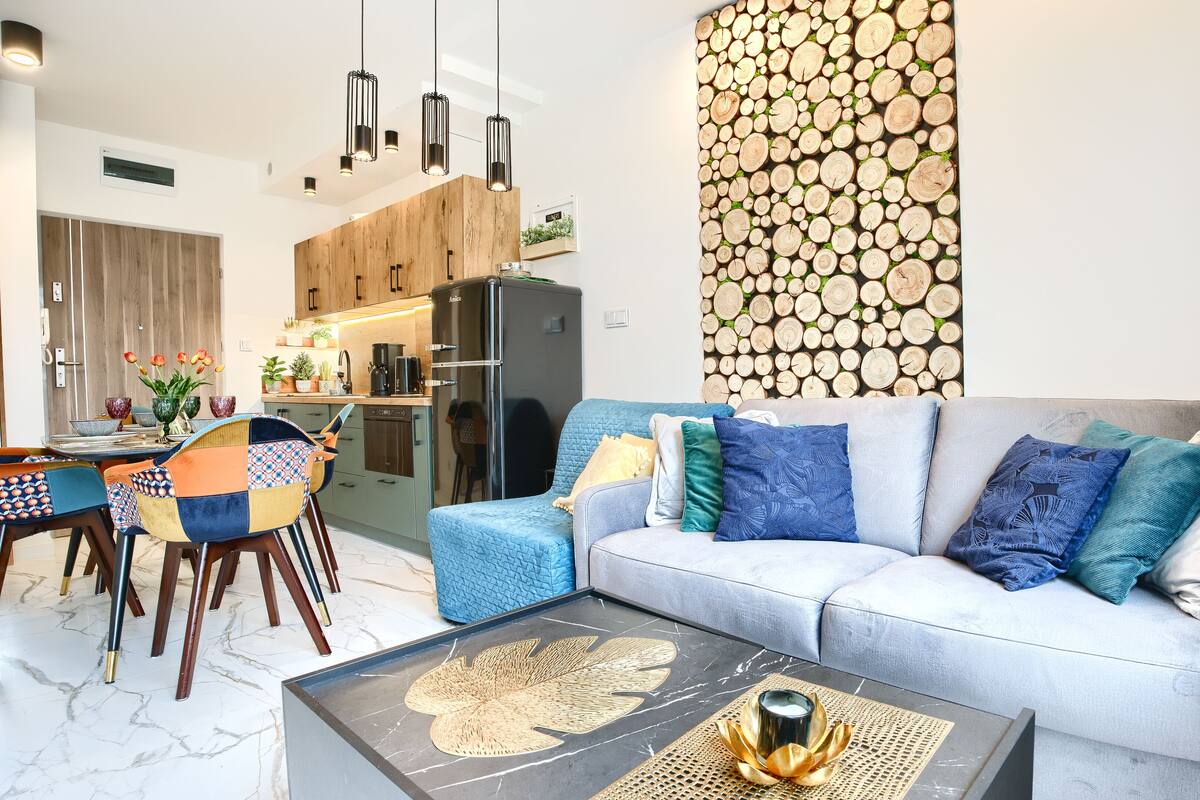 Ein modernes Wohnzimmer mit einem grauen Sofa, bunten Kissen, einem Couchtisch mit dekorativer Dekoration und einer Wand, die mit Holzscheiten gestapelt ist.
