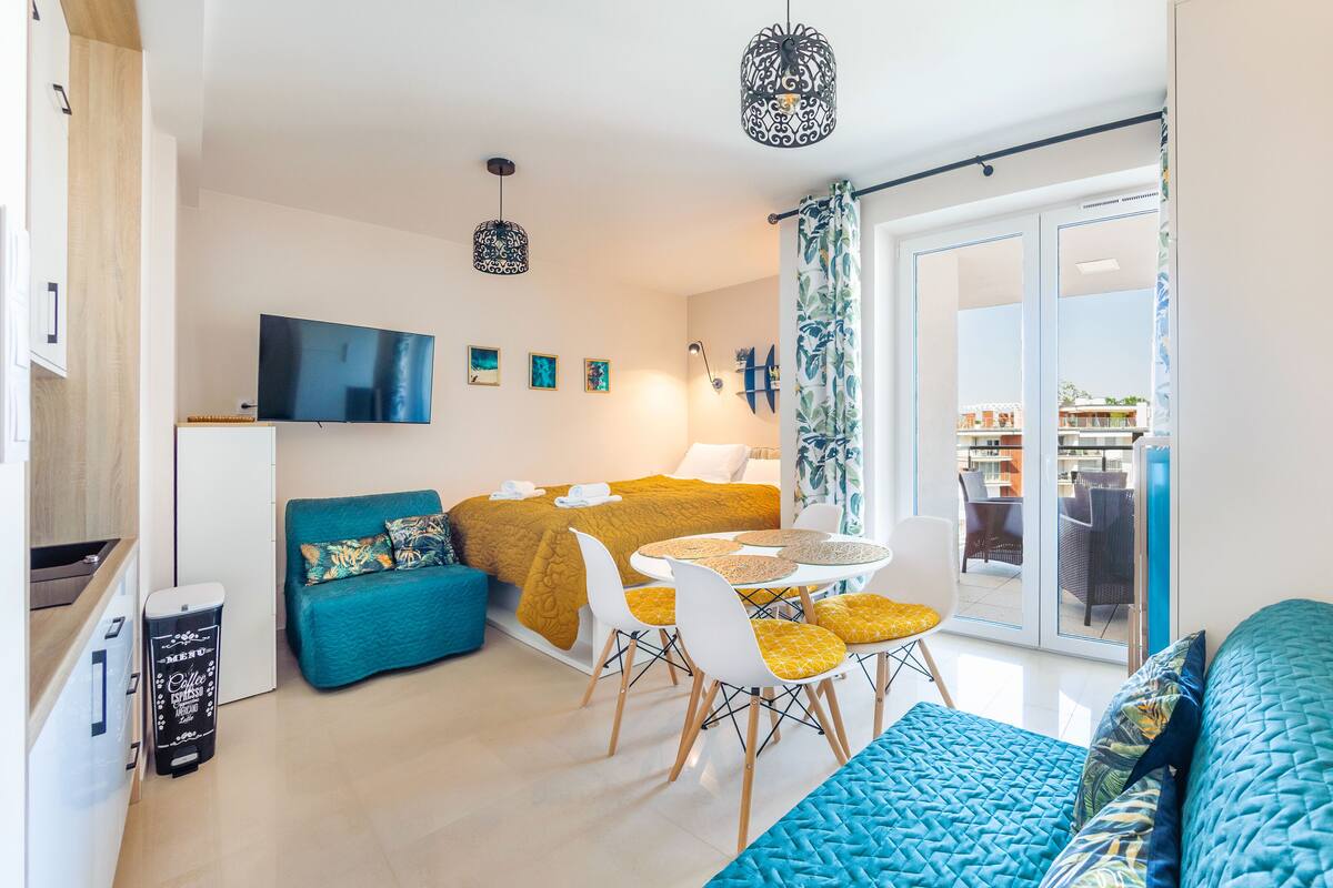 Ein modernes Wohnzimmer mit einer Couch, einem Esstisch mit Stühlen, einem an der Wand montierten Fernseher und einem Balkon mit Gartenmöbeln.