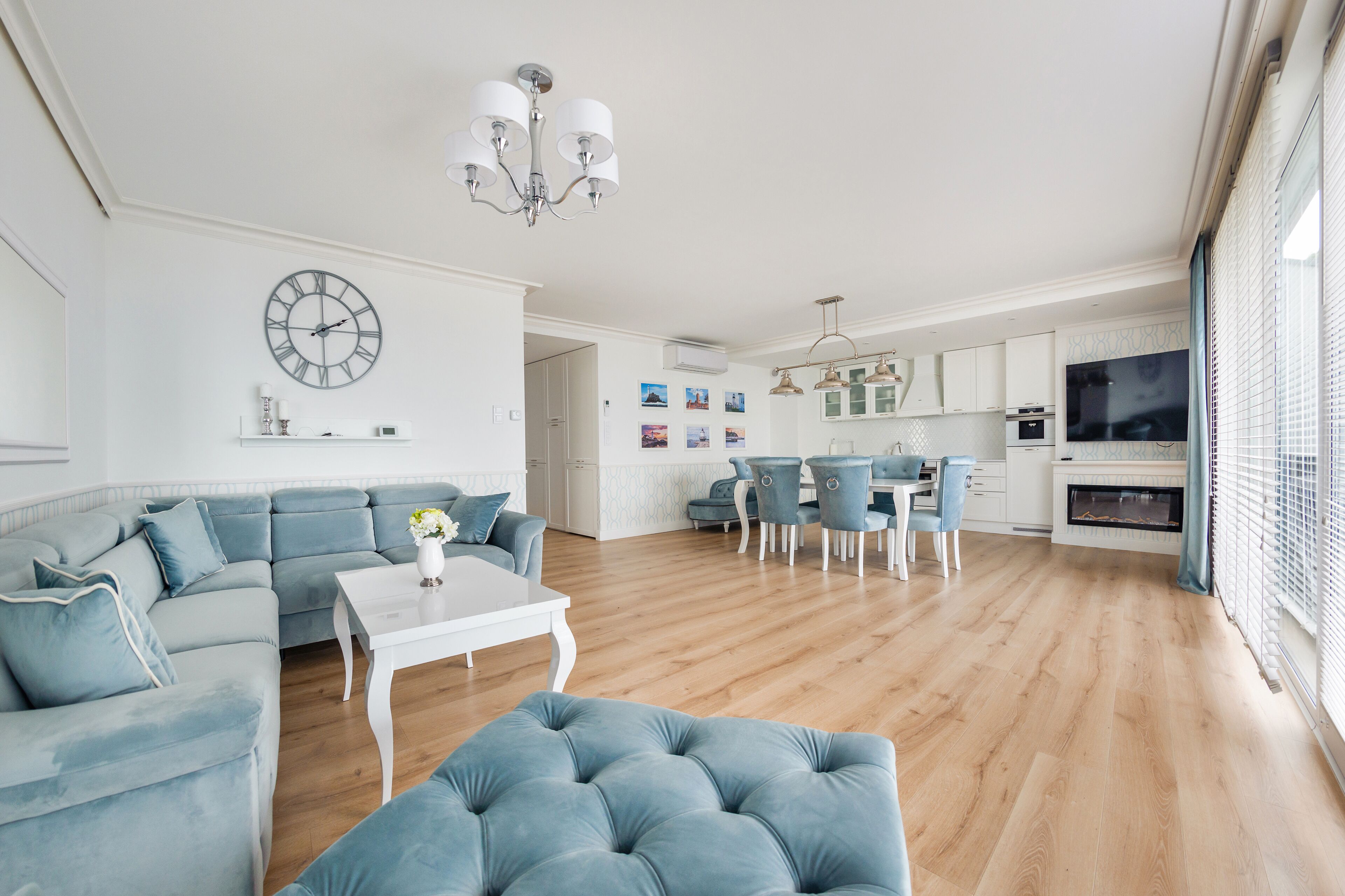 Una sala de estar moderna con un sofá seccional, una mesa de centro blanca y un reposapiés azul. Hay una zona de comedor con una mesa y sillas, y una cocina con armarios blancos y un ventanal grande.