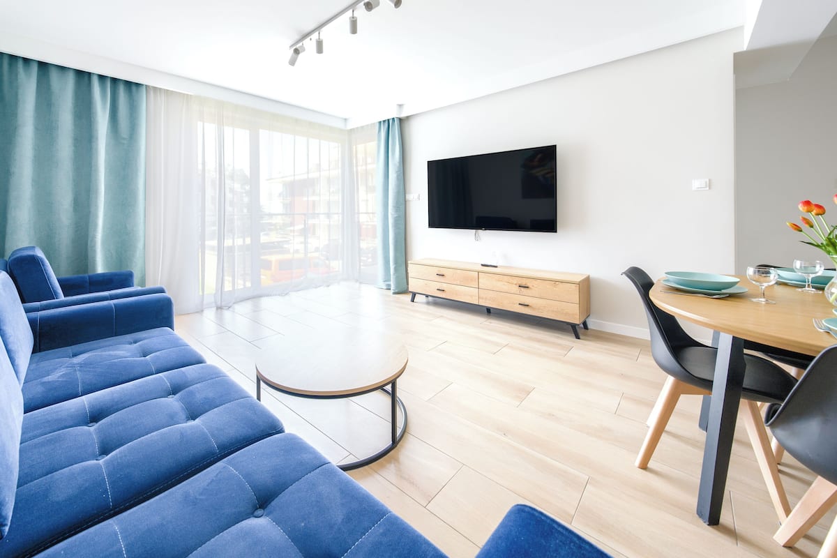 Ein modernes Wohnzimmer mit blauem Sofa, einem Esstisch mit Stühlen, einem an der Wand befestigten Flachbildfernseher und einem großen Fenster mit durchsichtigen Vorhängen.