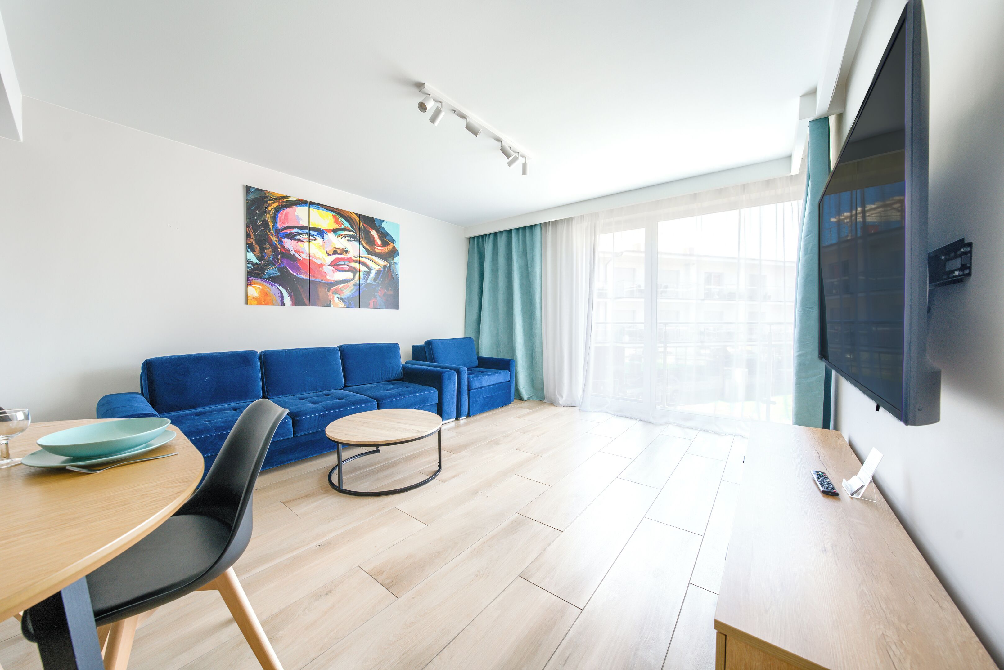 Ein modernes Wohnzimmer mit blauem Sofagarnitur, einem Esstisch mit Stühlen, einem flachbildfernseher an der Wand und einem großen Fenster mit Vorhängen.