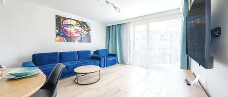 Ein modernes Wohnzimmer mit blauem Sofagarnitur, einem Esstisch mit Stühlen, einem flachbildfernseher an der Wand und einem großen Fenster mit Vorhängen.