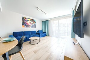 A modern living room with a blue sofa set, a dining table with chairs, a flat-screen TV mounted on the wall, and a large window with curtains.