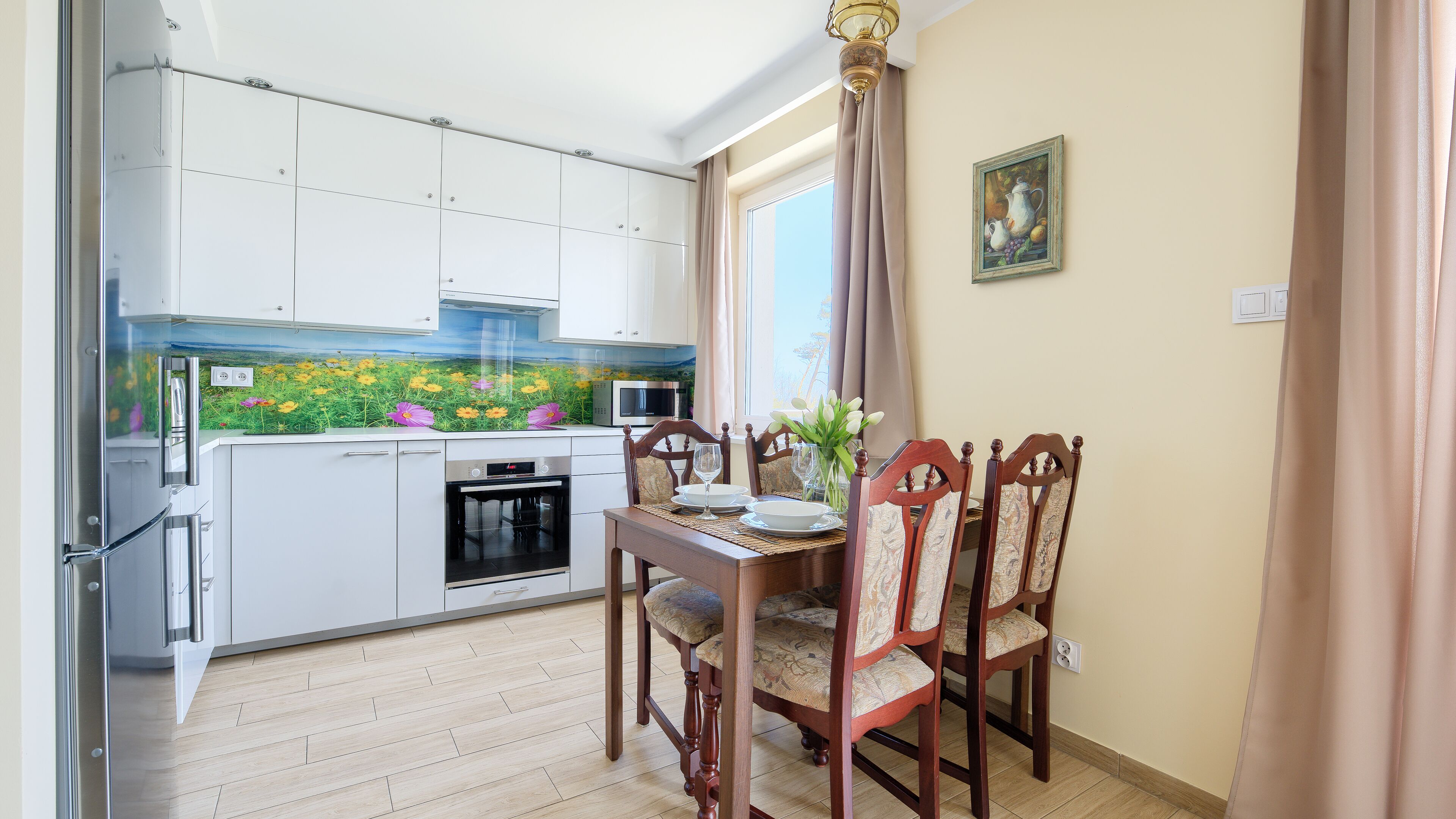 Cocina compacta con armarios blancos, un microondas y un horno pequeño. Hay una mesa de comedor con sillas y un florero con flores.
