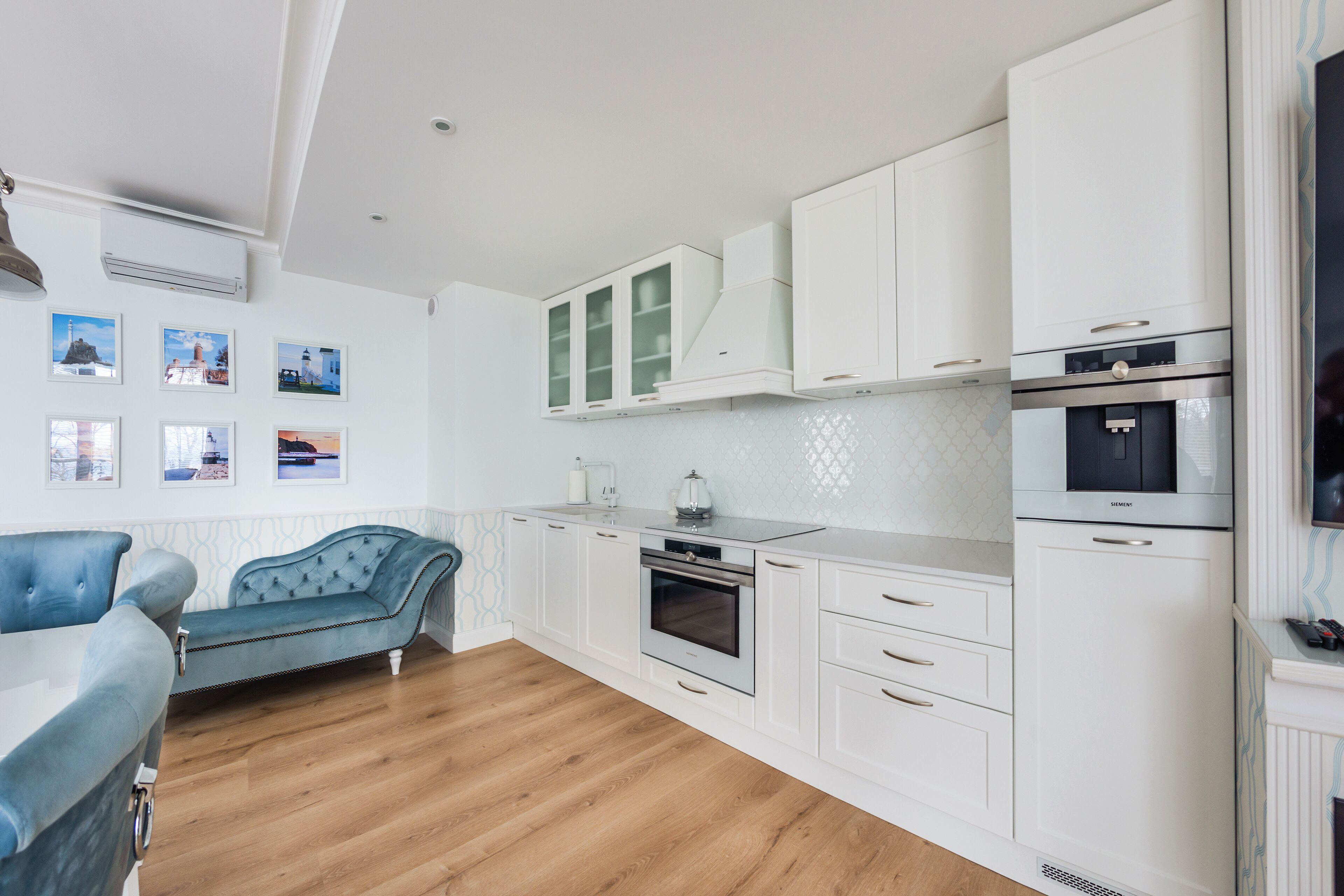Une cuisine moderne avec des meubles blancs, un four encastré et un four à micro-ondes. Le salon adjacent comprend un canapé bleu et une table à manger.