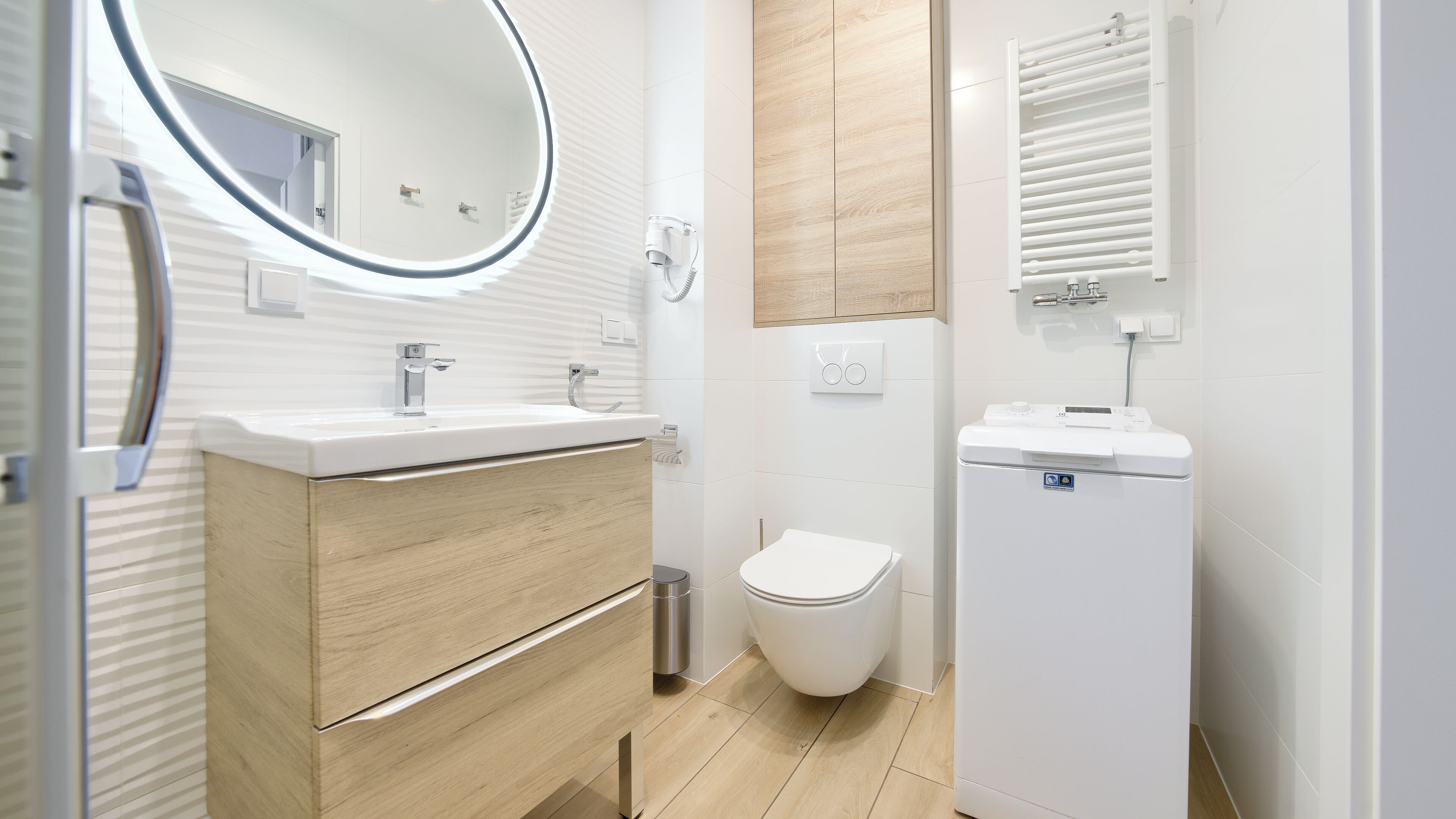 Een moderne badkamer met een houten wastafelmeubel, een ronde spiegel, een toilet en een apart wastafelunit.