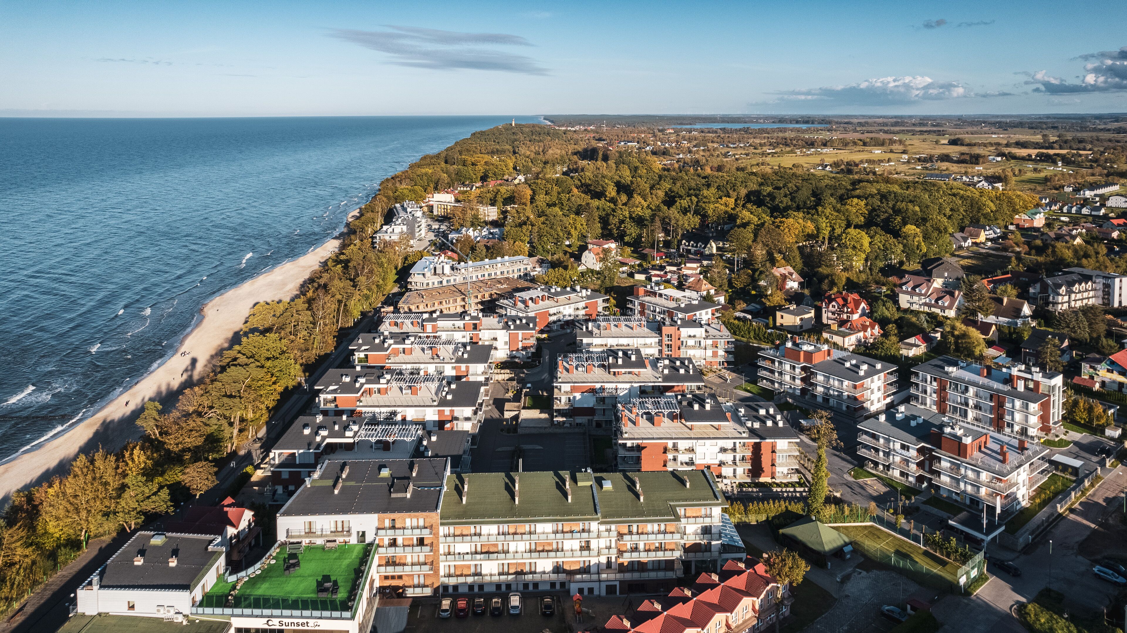An aerial view of a coastal residential area with a beach, houses, and a large building with a green roof.