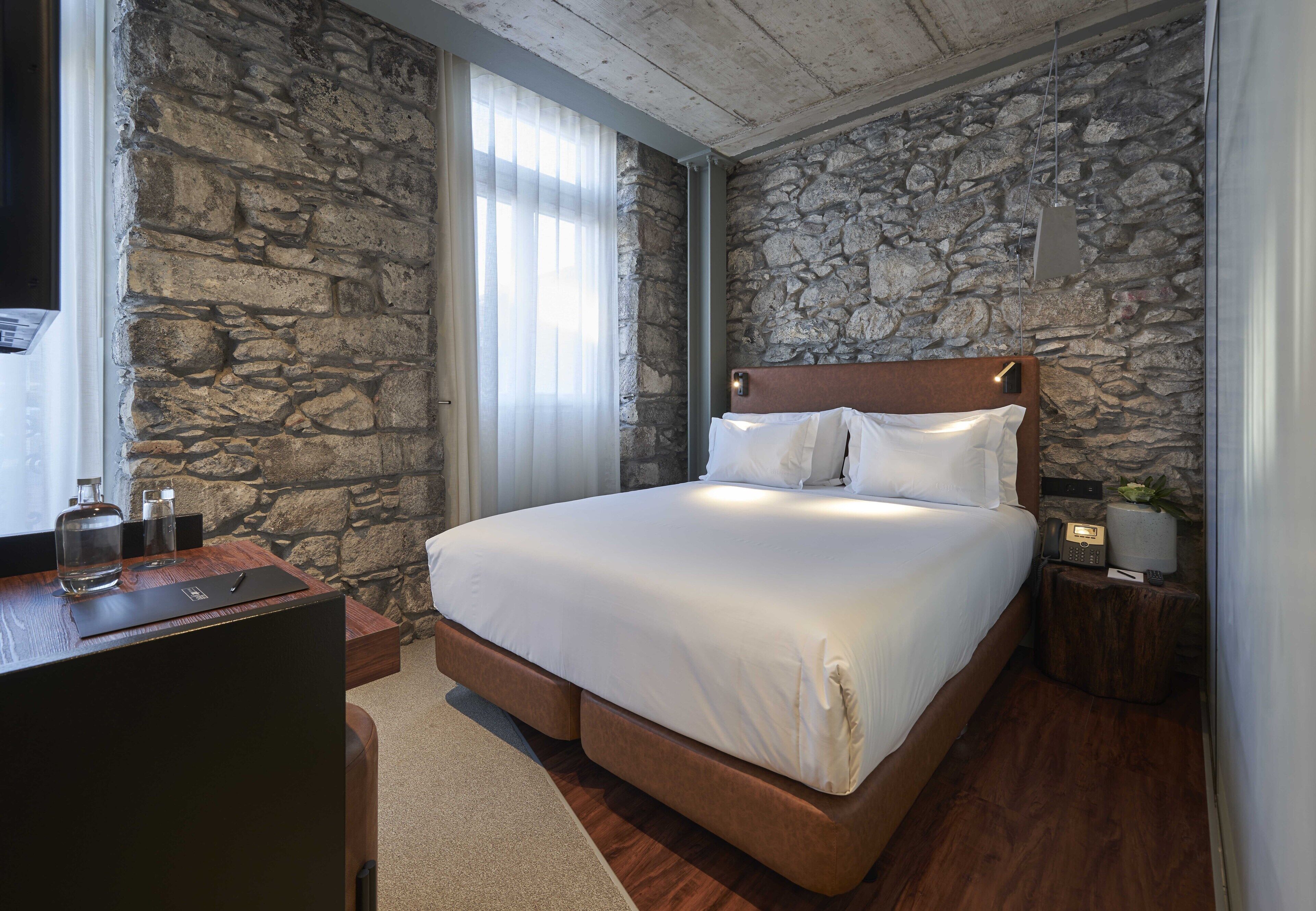 A bedroom with a stone wall, a wooden headboard, a bed with white linens, a bedside table with a lamp, and a small nightstand with a radio.