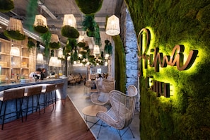 A modern restaurant interior with hanging green plants, wicker furniture, and a neon sign on the wall. - Hotel Caju (Funchal)