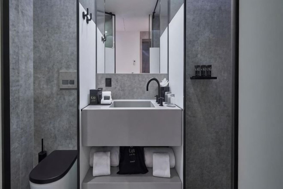 A modern bathroom with a white sink, black faucet, and a mirror above it. There are towels and a black object on the counter.