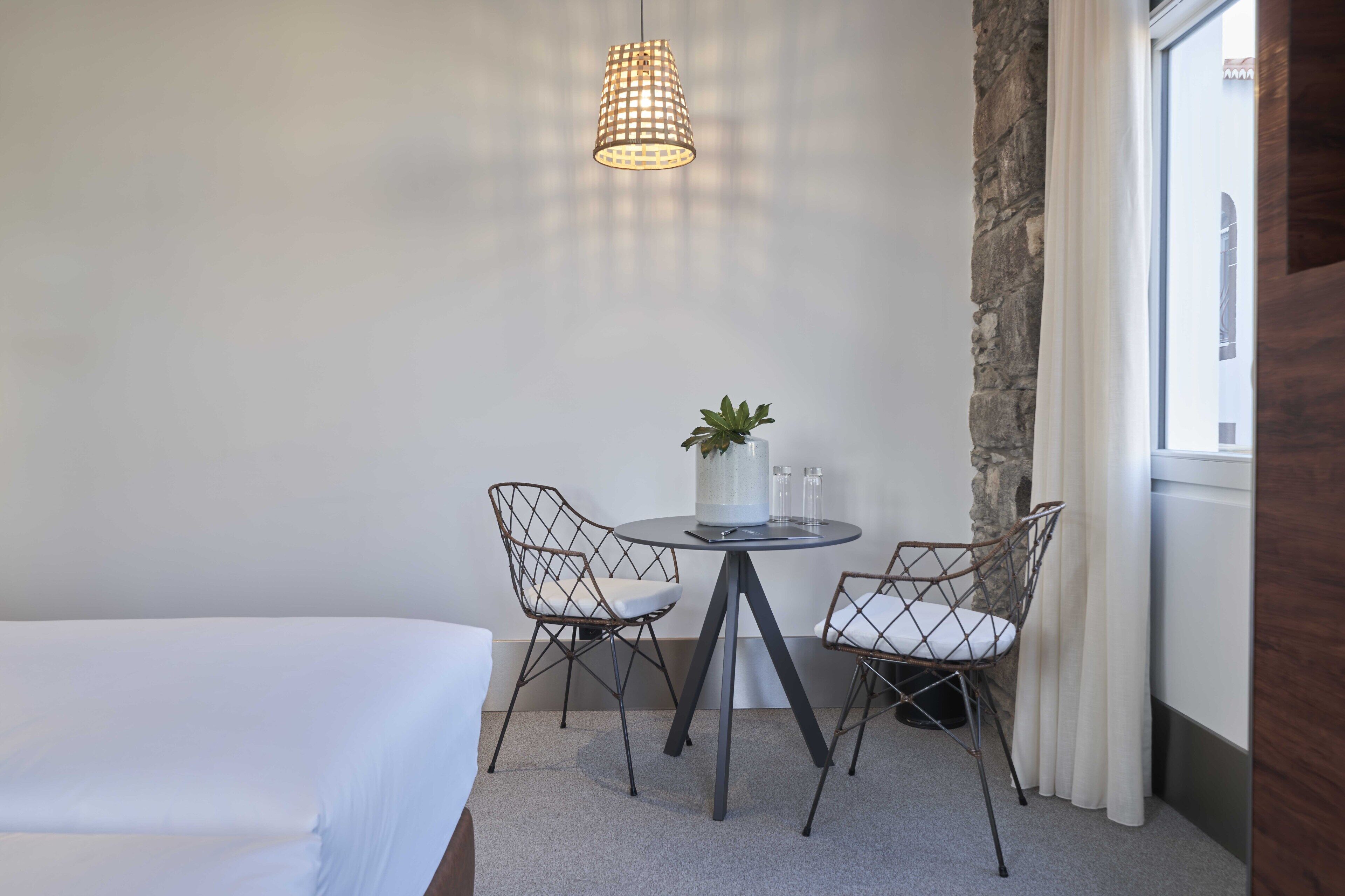 Une chambre d’hôtel avec un lit, une petite table ronde et deux chaises, une plante en pot et un luminaire suspendu.