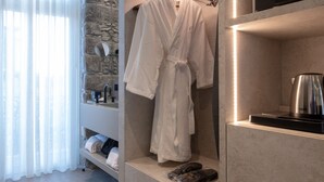 A modern bathroom with a stone wall, a robe hanging on a rack, a kettle, and neatly folded towels on a shelf. - Hotel Caju (Funchal)