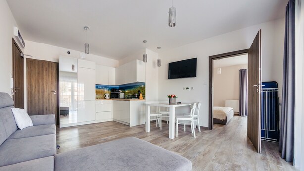 A modern living room with a grey sofa, a dining area with a table and chairs, a kitchen with white cabinets, and a TV mounted on the wall. - Apartamenty Sun & Snow Piec Morz (Ustronie Morskie)