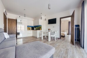 A modern living room with a grey sofa, a dining area with a table and chairs, a kitchen with white cabinets, and a TV mounted on the wall. - Apartamenty Sun & Snow Piec Morz (Ustronie Morskie)