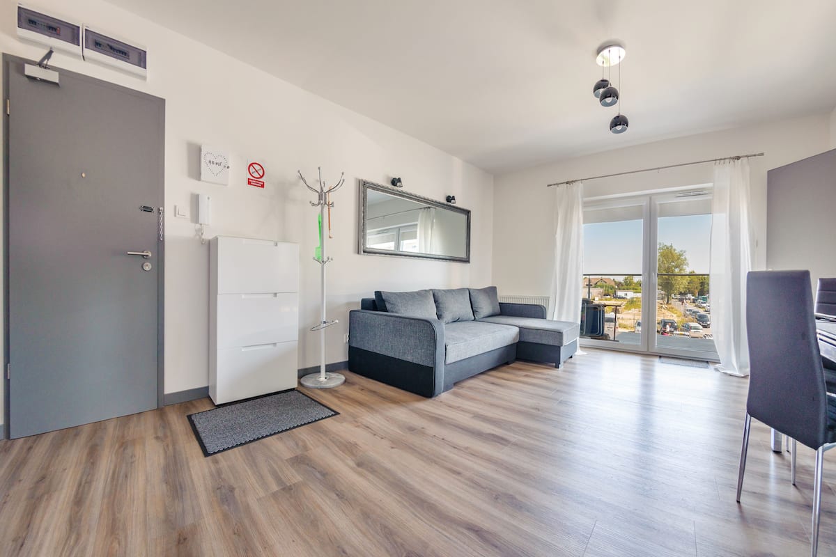 A modern living room with a grey sofa, a flat-screen TV, and a large window offering a view of the outdoors.
