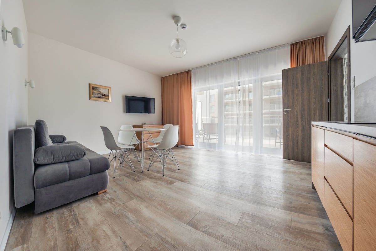 A modern living room with a grey sofa, a dining table with chairs, a flat-screen TV, and a large window with curtains.