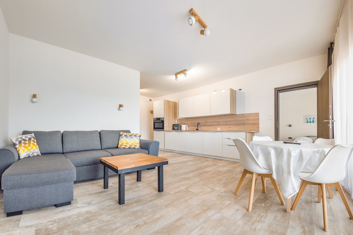 A modern living room with a grey sofa, a wooden coffee table, and a dining area with white chairs and a table.