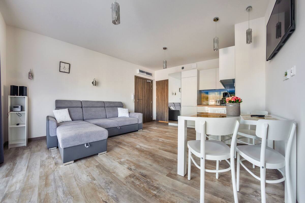 A modern living room with a grey sofa, a dining table with white chairs, and a kitchen area in the background.