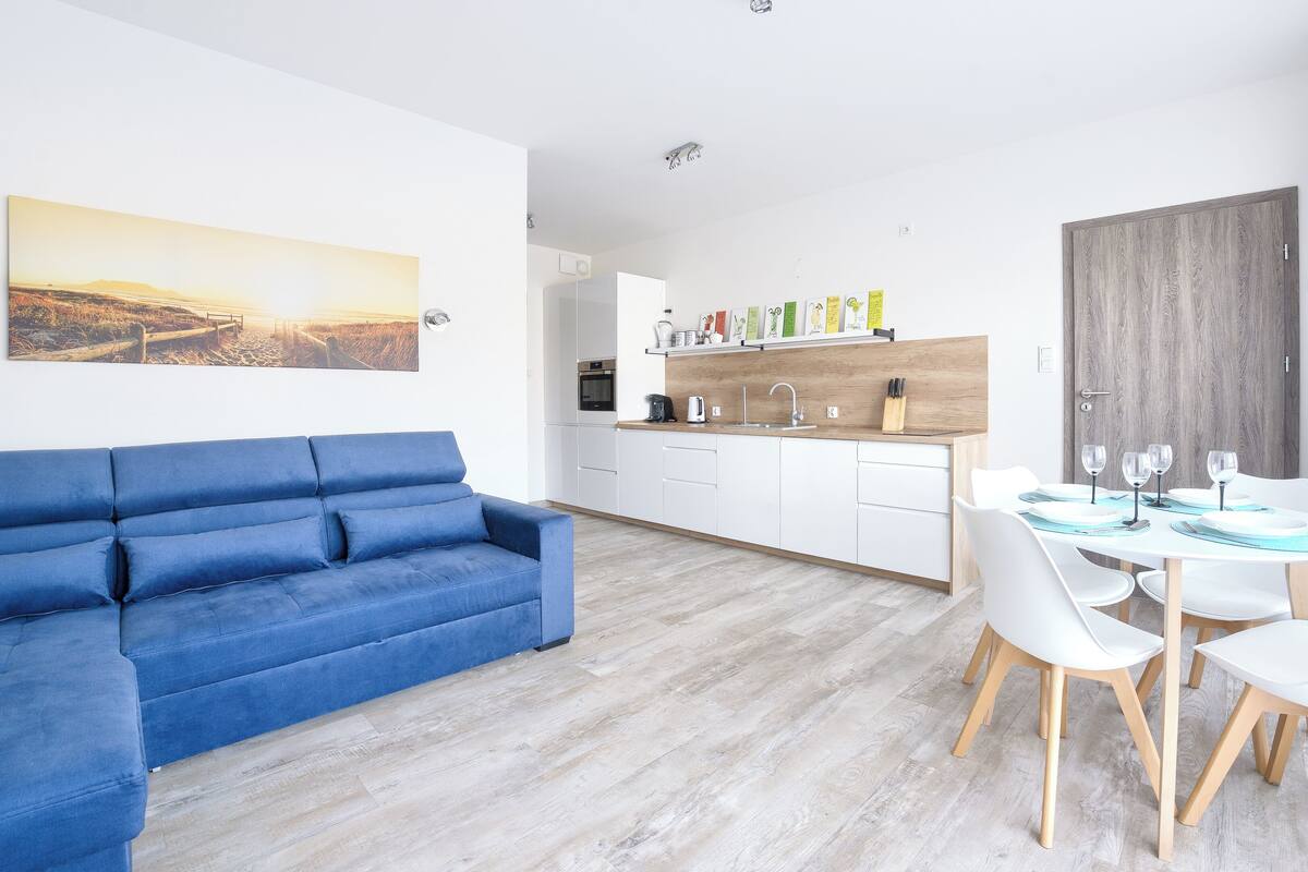 A modern living room with a blue sofa, a dining area with white chairs and a table, and a kitchen with white cabinets and a wooden countertop.