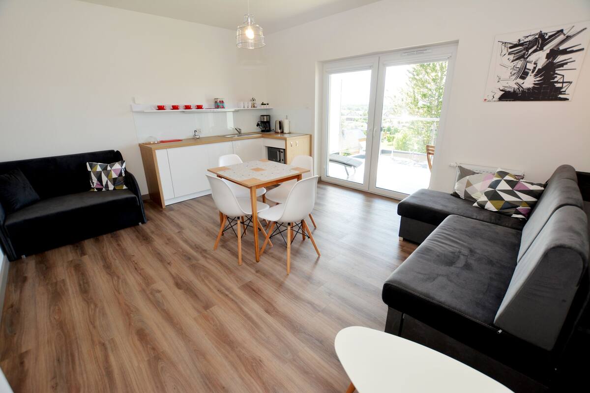 A modern living room with a black sofa, a white dining table with chairs, and a kitchen area with a coffee machine.